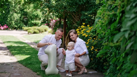little baby boy stands near the balloon of number one shape. happy smiling parents sit beside their child outdoors in summer.