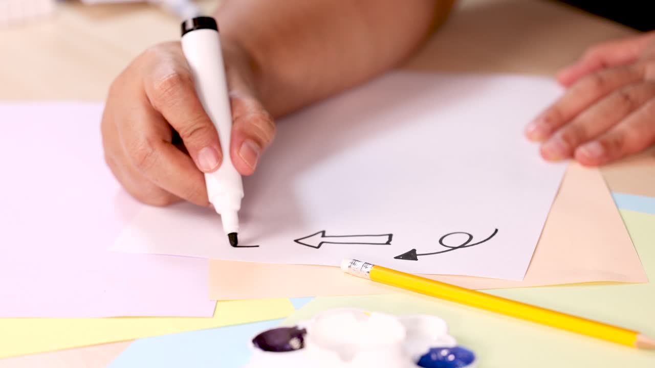 Person draws black arrow and swirl on paper using marker, overhead view, bright lighting