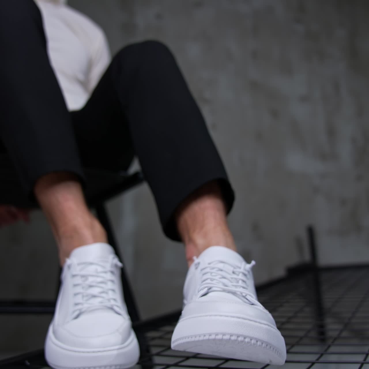 Male sitting on the chair demonstrating modern sport footwear. Unrecognized man in white shirt and black trousers presents white sneakers in front of camera. Close up. Blurred backdrop