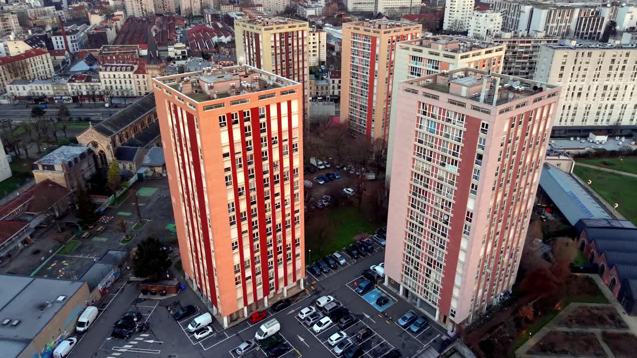 Foto aérea com drone orbitando dois edifícios residenciais altos, laranja e brancos, em um subúrbio de Paris, com uma igreja visível no plano de fundo