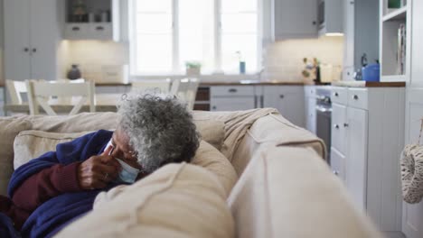 sick senior african american woman covered with blanket sneezing while lying on the couch at home