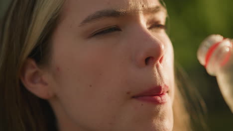 close-up of woman drinking orange juice from a plastic bottle outdoors, reflecting satisfaction, sunlight shining on her face and bottle, with a soft glow of light and greenery in the background