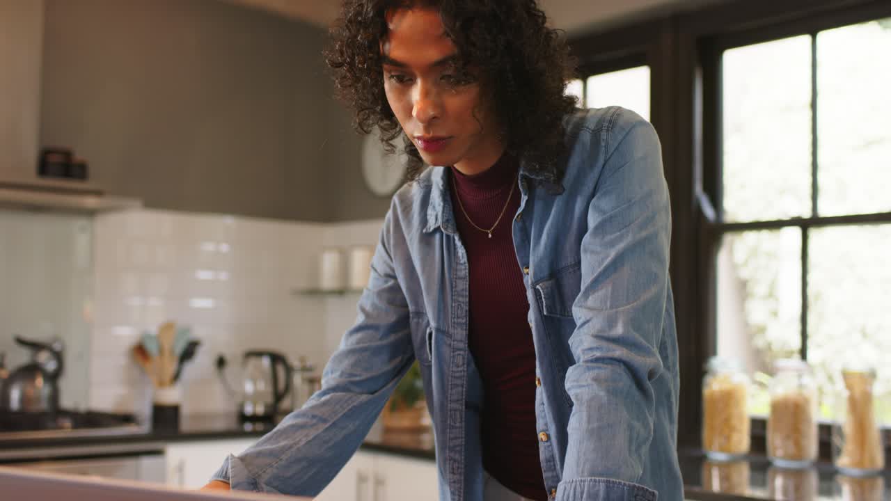 Non-binary person leaning at kitchen counter reviewing laptop reaching mug sipping to stay focused