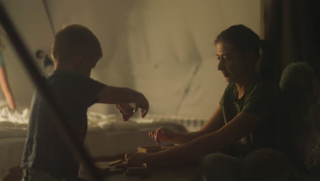 young mother and little son laying out wooden set for board game jenga on small portable table in front of transparent window. family board game night