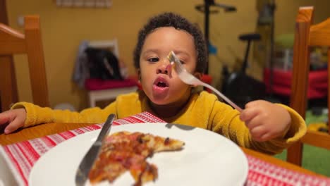 cute and very expressive two-year-old black baby eats pizza by himself with a fork at home