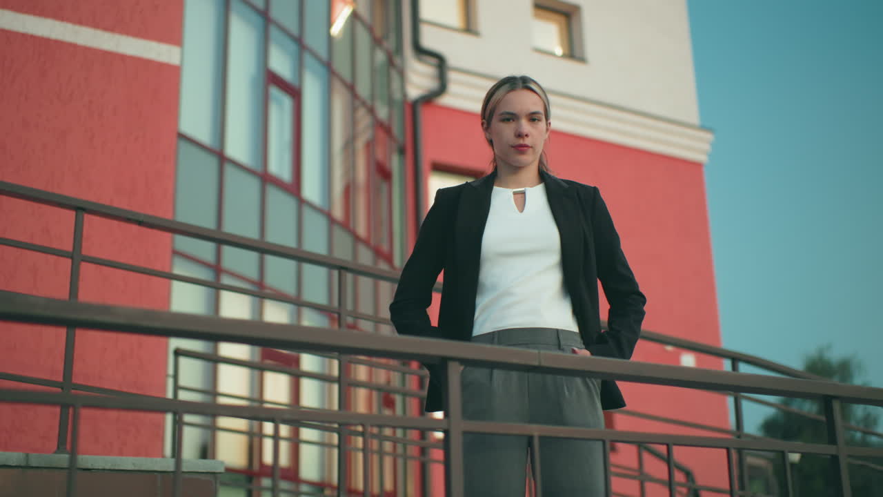 Elegant woman in black blazer and white blouse stands confidently with hands in pockets in front of residential building, iron railing and street light visible, tree in background