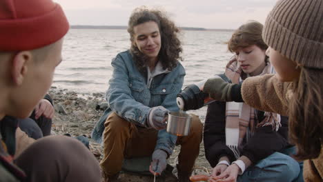 group of teenage friends sitting around the bonfire while they serving tea in cups with a thermos on the seashore