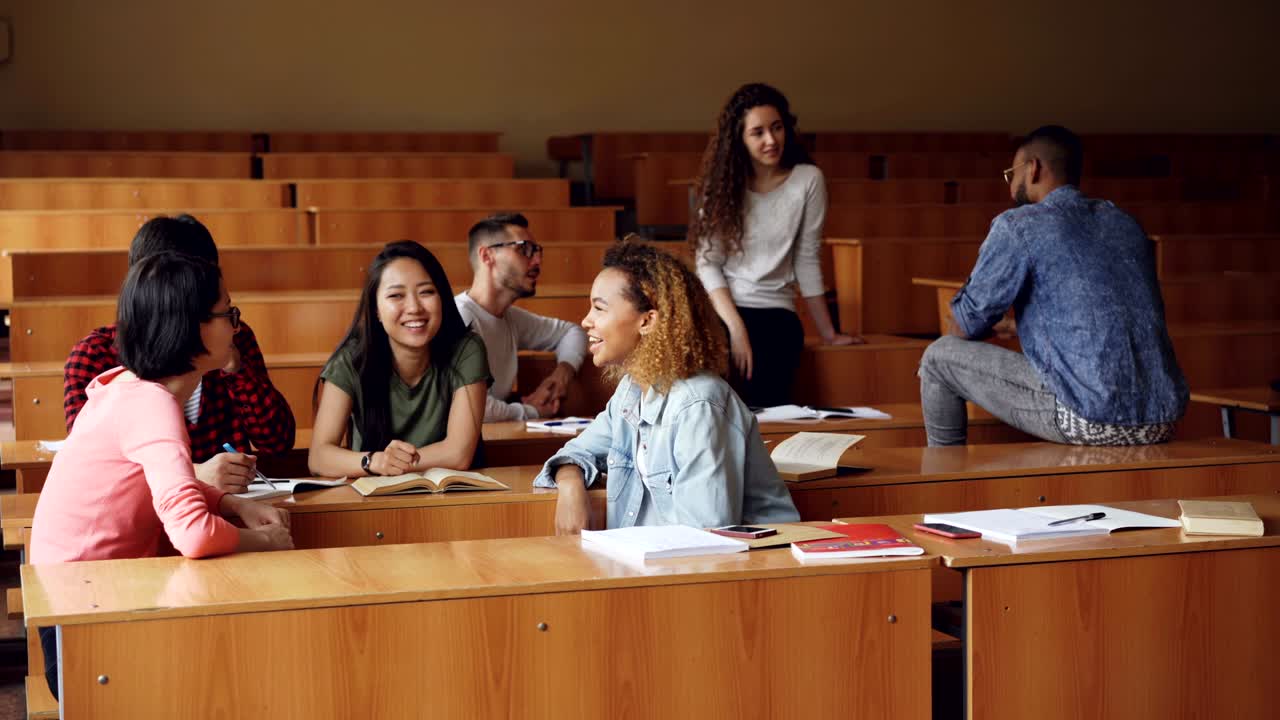 학생들의 다민족 그룹은 휴식 시간과 의사소통을 즐기며 휴식을 취하고 대화하고 있습니다. 나무 테이블, 매력적인 사람들 및 노트북이 보입니다.