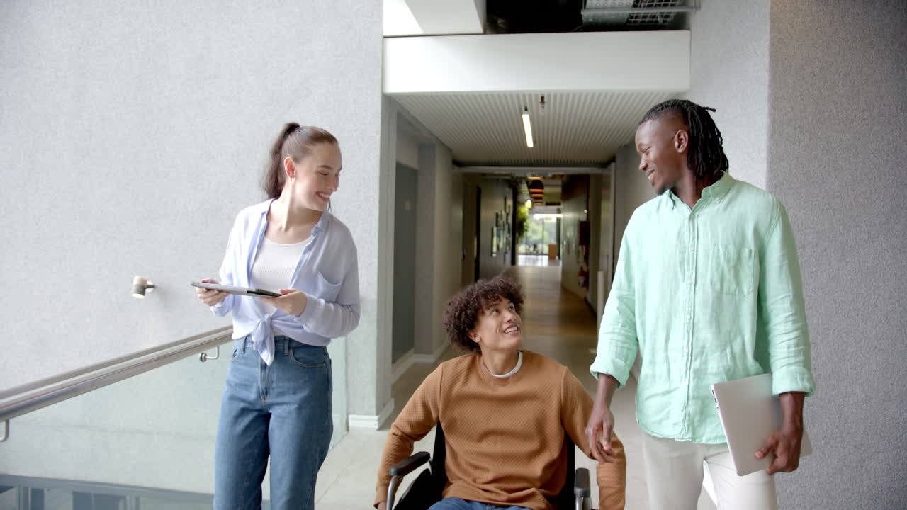 Walking with man in wheelchair, diverse colleagues discussing work using tablet and laptop