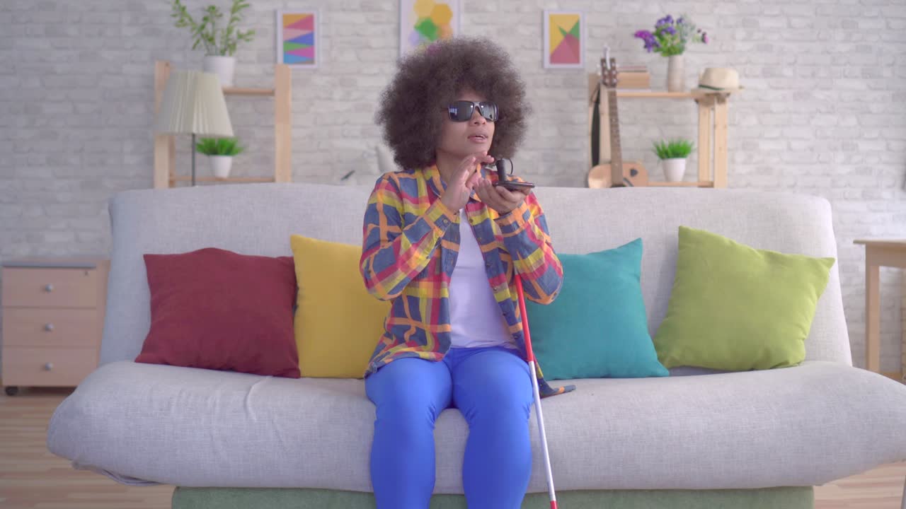 mujer afroamericana con un peinado afro con discapacidad visual en gafas usa asistente de voz en su teléfono inteligente sentada en el sofá