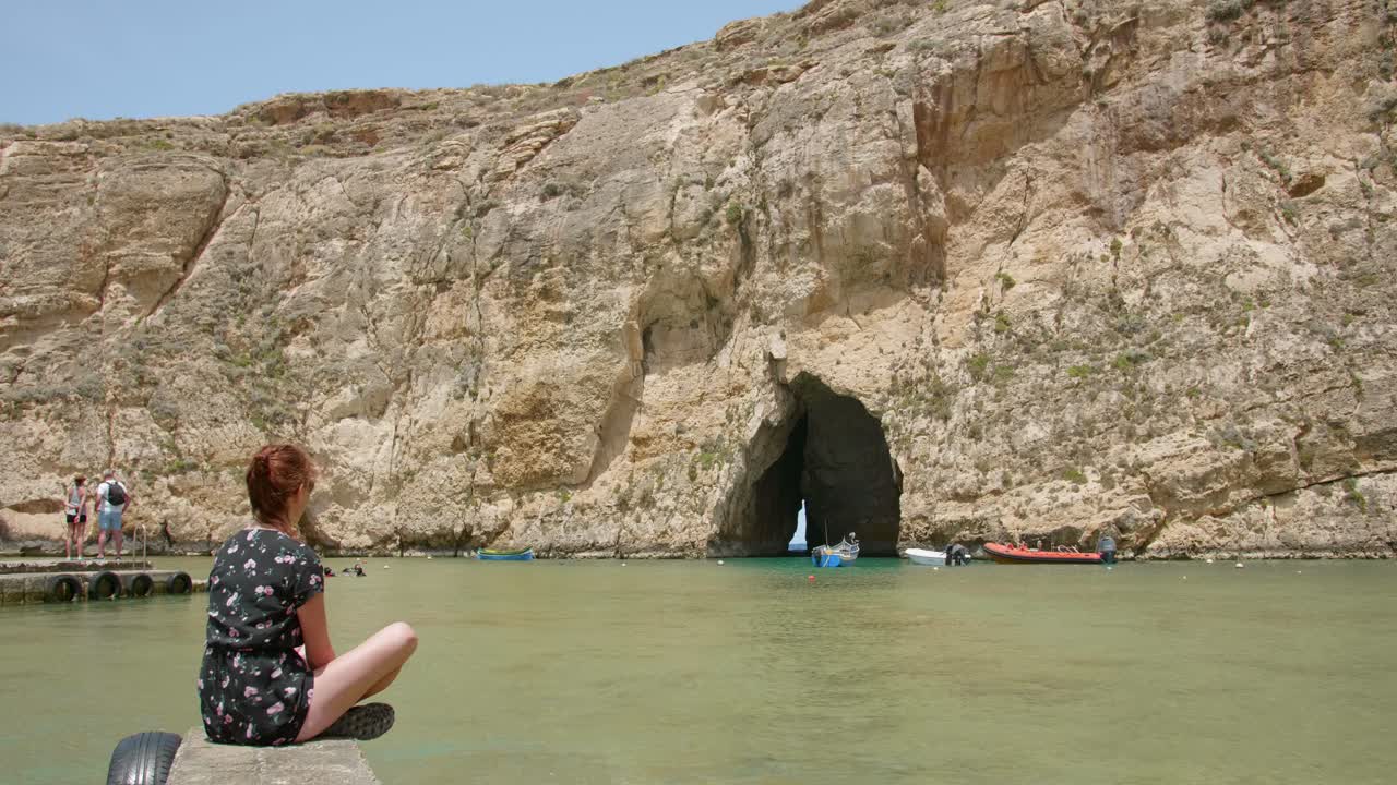 mujer vestida sentada en una roca mientras observa el mar interior y los barcos que pasan por una cueva en la isla de gozo en malta en un día soleado de verano