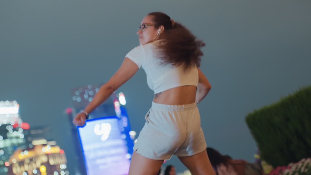 Donna che balla di notte su un tetto, ascoltando musica con gli auricolari, braccia fluide e volteggi energici, smartphone in mano. Gesti in primo piano ed espressione spontanea, con lo skyline sfocato e le luci al neon che creano un'atmosfera suggestiva e gioiosa.