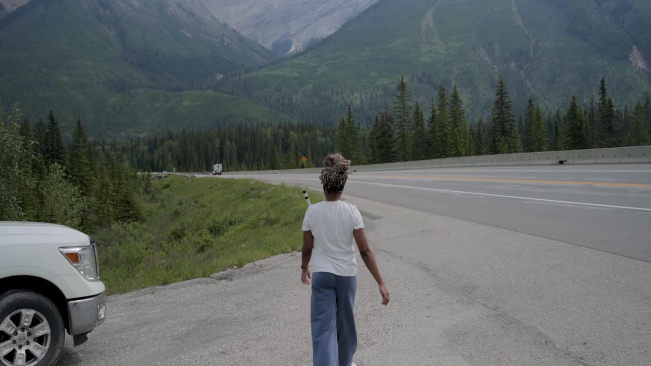 Black lady walking along the side of a highway with the mountains ranges of Yoho National Park in front of her in Yoho National Park