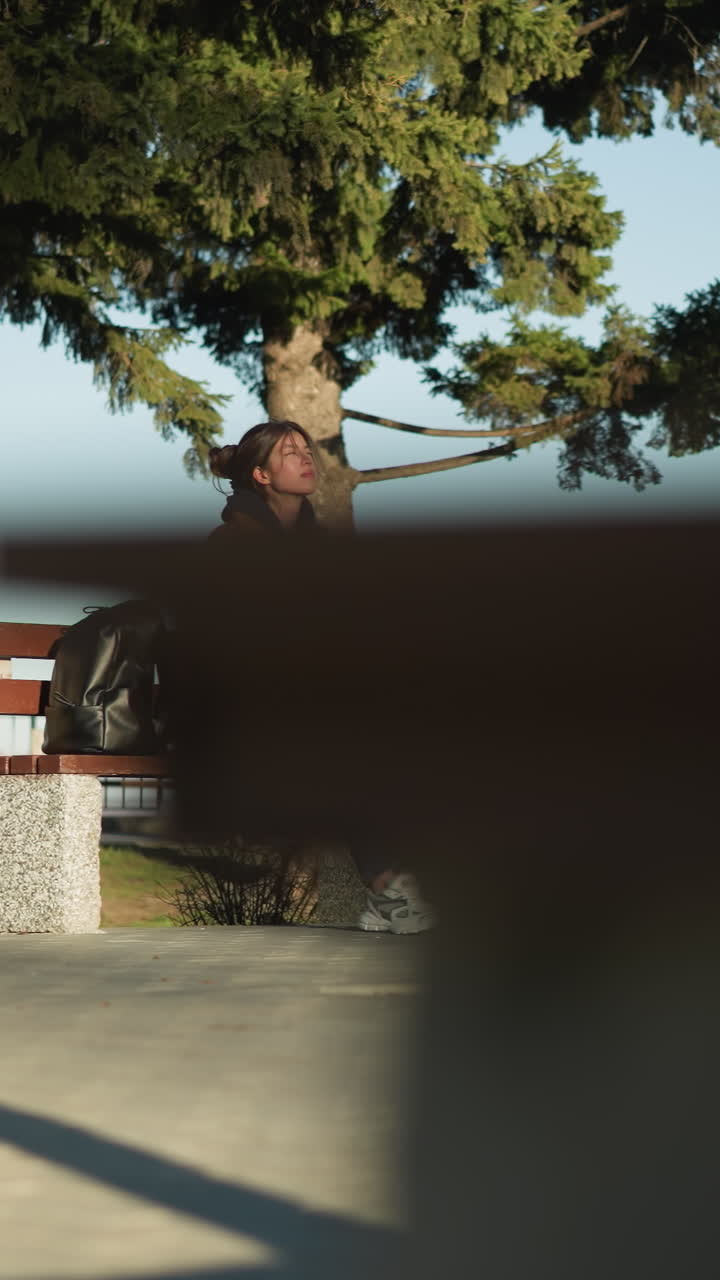 한 소녀가 도시 공원의 벤치에 혼자 앉아 슬프고 생각에 빠진 모습을 보이고, 갈색 코트, 파란색 청바지, 색 신발을 입고, 에 검은색 배을 고 있다.