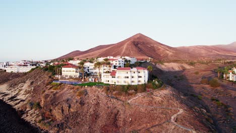 Coastal-resort-town-nestled-against-a-hill-with-a-prominent-mountain-backdrop,-under-clear-skies,-aerial-view