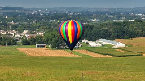 Toma-Panorámica-Aérea-De-Un-Colorido-Globo-Aerostático-Volando-Sobre-Campos-Verdes-En-Las-Afueras-De-Una-Ciudad-Americana