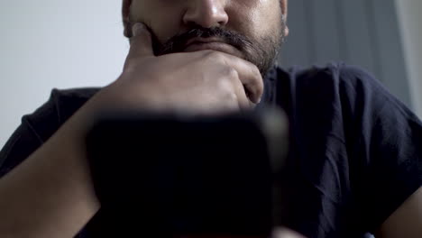 a close-up shot of a middle-aged adult male, who is touching his beard while reading something on his cellphone