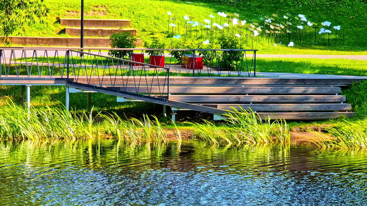 Wooden Pedestrian Bridge in Park Above Water with Green Grass and Seating Area