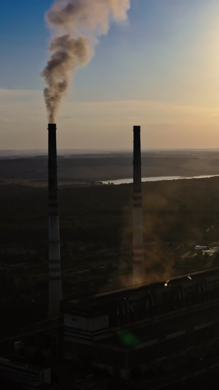 Dark silhouette of a factory at sunset. Manufacture releases harmful smoke into the air. Nature in danger. Industrial plant near the river in the evening. Vertical video
