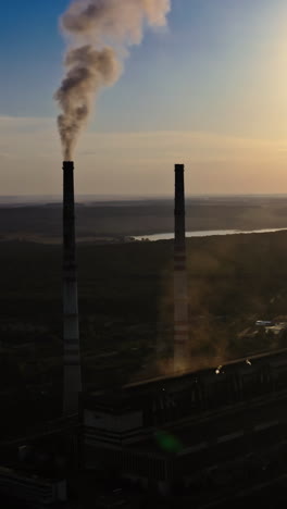 dark silhouette of a factory at sunset. manufacture releases harmful smoke into the air. nature in danger. industrial plant near the river in the evening. vertical video