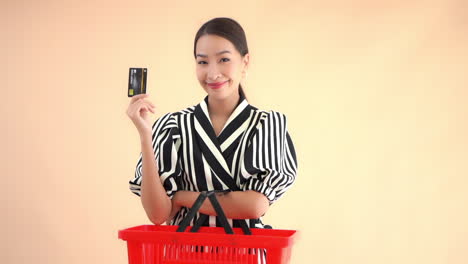 Asian-woman-with-red-shopping-basket-on-arm-and-holding-credit-card-smiles-at-camera