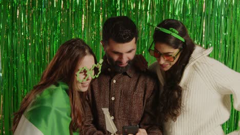 studio shot of friends dressing up with irish novelties and props posing for selfie celebrating st patrick's day against green tinsel background 2