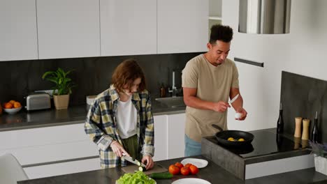 Happy-young-man-with-Black-skin-color-brunette-in-a-beige-T-shirt-prepares-scrambled-eggs-while-his-mature-young-girl-in-a-plaid-shirt-prepares-a-salad-while-they-are-preparing-their-breakfast-in-the-morning