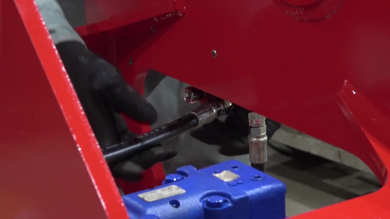 Technician connecting a hydraulic hose fitting to a blue control valve block on a red heavy construction machinery frame