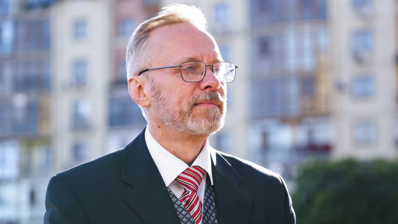 Close-up portrait of a senior man with grey hair with beard sitting on the city background. Old businessman in eyeglasses straighten his tie and looks to camera.