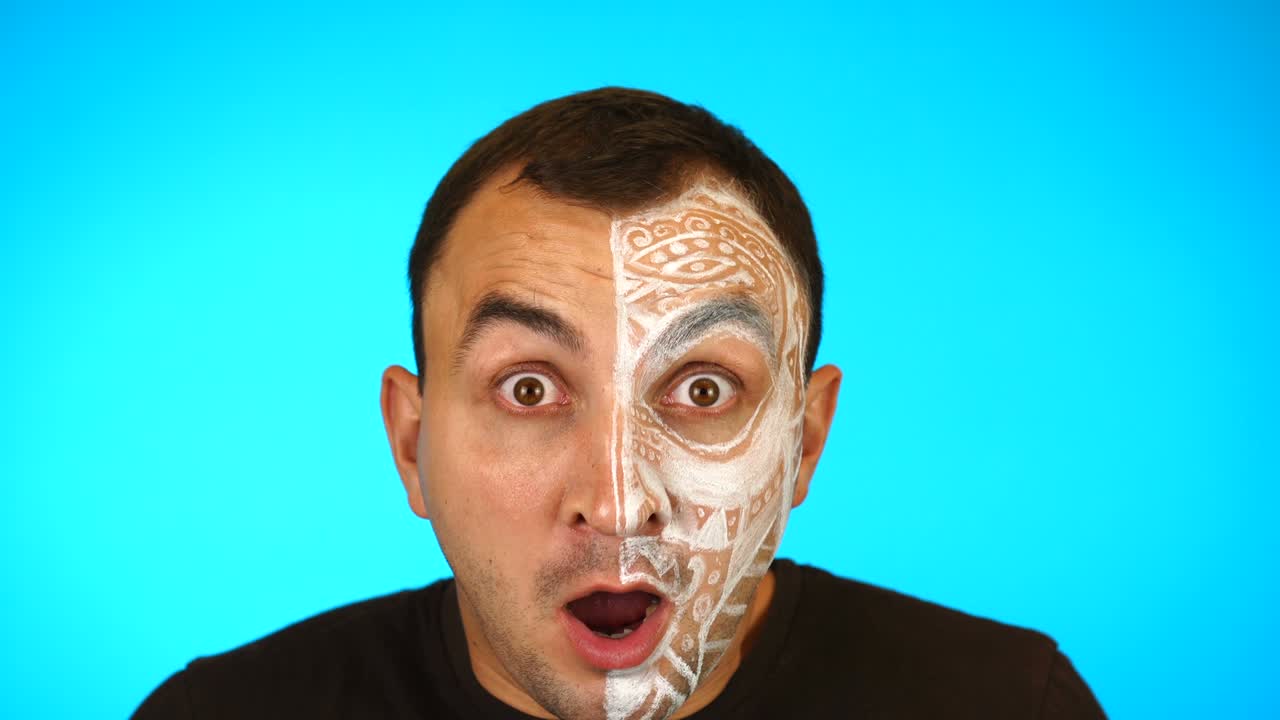 얼굴이 칠해진 남자는 놀랐거나 충격을 받았고 파란색 배경에 카메라를 보고 있습니다.