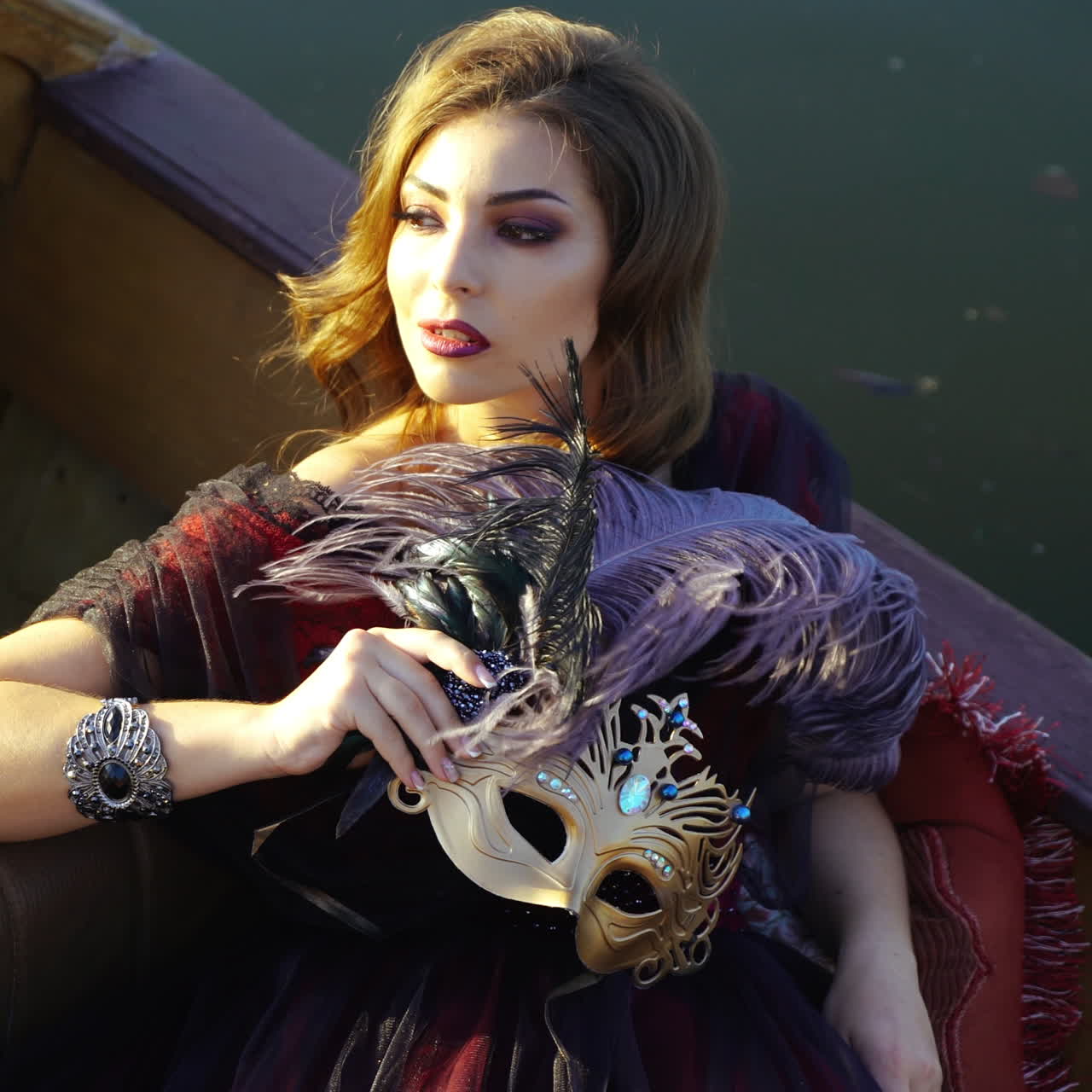 Portrait of a dreamy young woman with bright makeup sitting in a boat and holding masquerade mask. Attractive girl floating in gondola over the river. Close-up