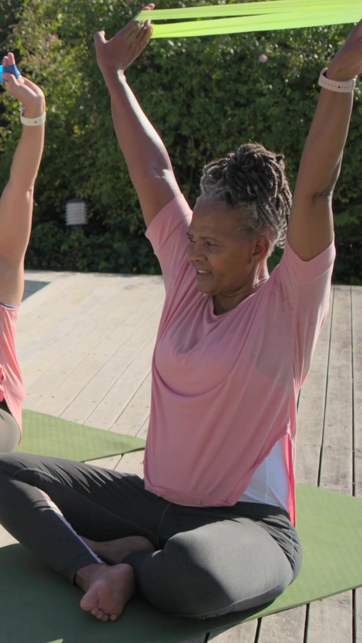 Video verticale: Donna a destra che tira una fascia verde sopra la testa dal petto, su una terrazza in rosa, per fare stretching.