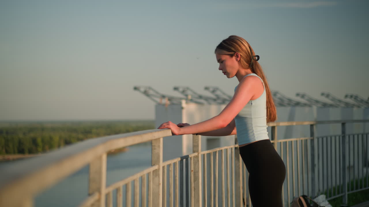 젊은 여자가 철의 레일링을 고, 따뜻한 빛 아래 멀리 바라보는 동안 손을 쉬고, 오른쪽 다리가 앞으로 나아가고, 평온한 포즈, 배경은 멀리 있는 초록색과 현대적인 구조물입니다.