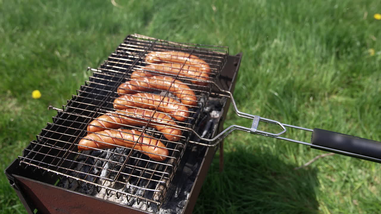 Grilling food for picnic. Delicious hot dog sausages frying in a grill grate in smoke on green nature background. Barbeque in summer.