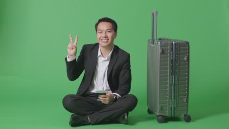 짐과 여권을 가진 아시아 사업가 여행자의 몸이 가득 차 있으며, 녹색 스크린 배경 스튜디오에 앉아있는 동안 미소 짓고 평화의 몸짓을 보여줍니다.