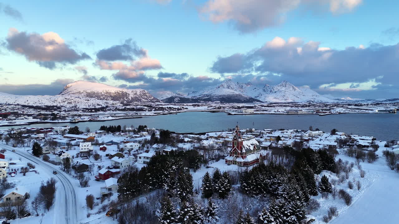 Cinematic Aerial view of Buksnes Church in Lofoten, Norway, surrounded by snow-covered landscape, red wooden architecture, fjord coastline and dramatic winter mountains at sunset