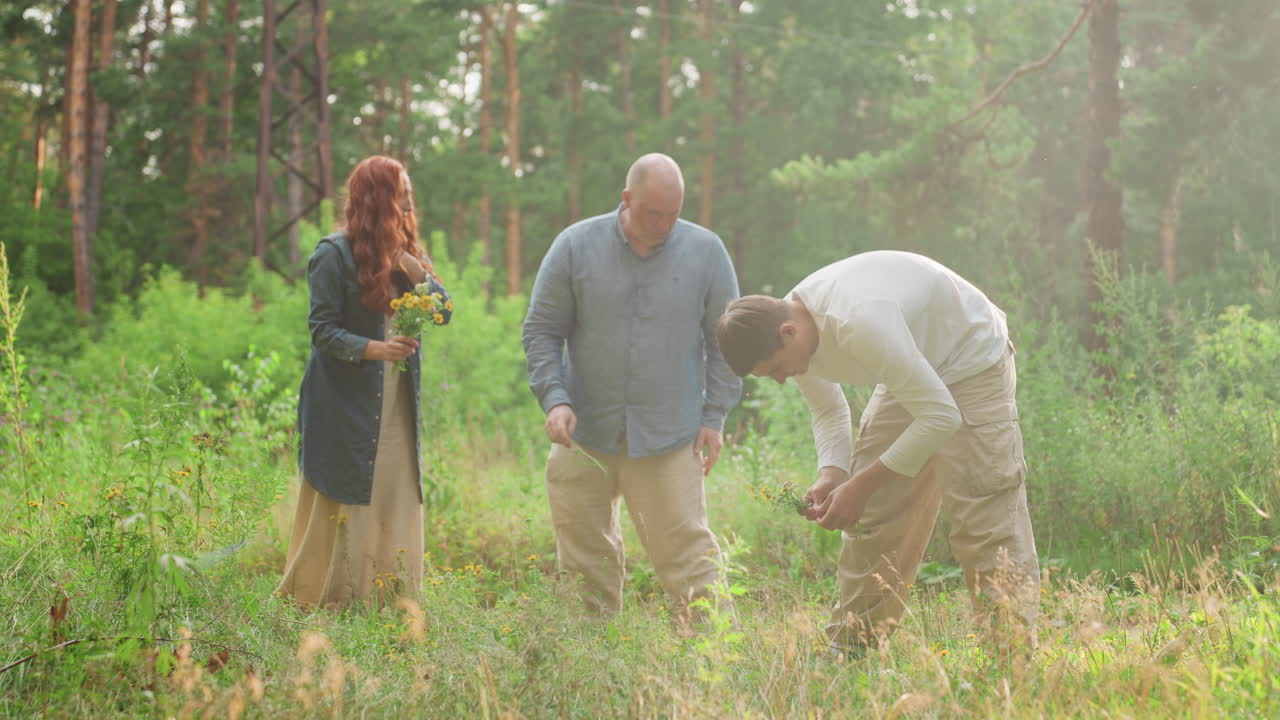 Picnic family gathering wildflowers in sunny green forest, enjoying peaceful outdoor moment, parents and son picking fresh bouquets surrounded by nature, showing unity, and calm lifestyle