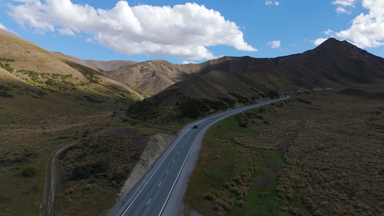 vista panorámica aérea del paso de lindis en nueva zelanda, que muestra una carretera sinuosa que atraviesa un terreno árido y montañoso bajo un cielo azul vívido salpicado de nubes.