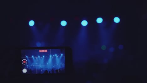 a close-up shot of a man's hand is taking video for a music band performing on stage with the neon light at the night club.