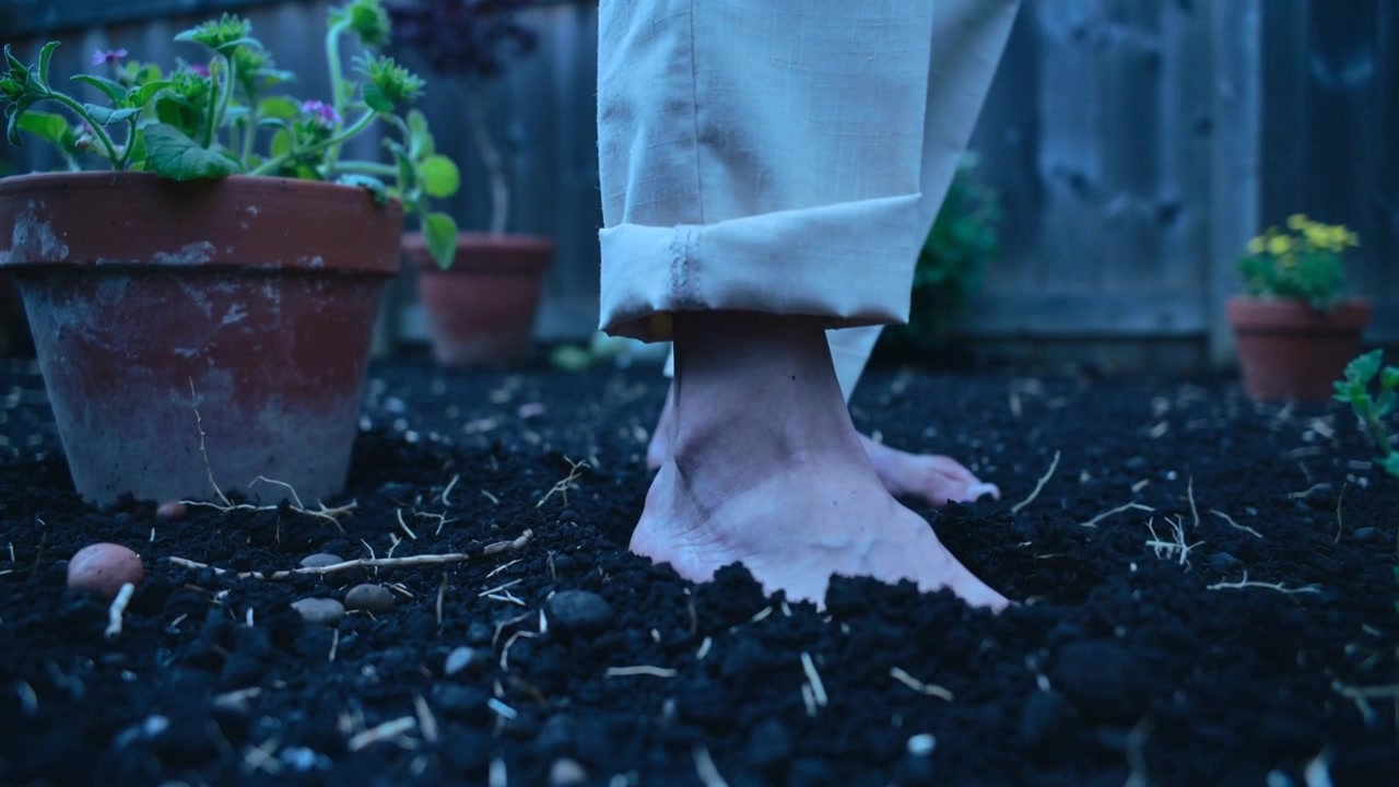 Stepping bare feet and ankles pressing into backyard soil near terracotta pots, with rolled cuffs