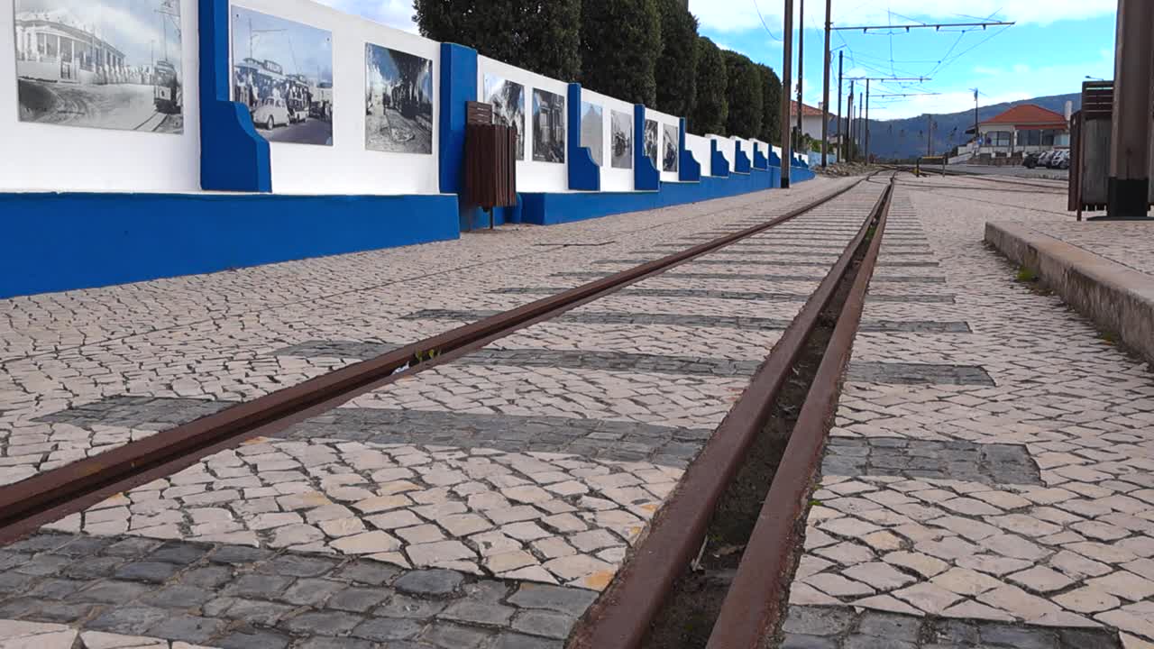 Plano cenital cinematográfico a lo largo de las vías oxidadas del tranvía de Sintra incrustadas en el pavimento de adoquines de calçada portuguesa tradicional en Azenhas do Mar en un día nublado sin gente