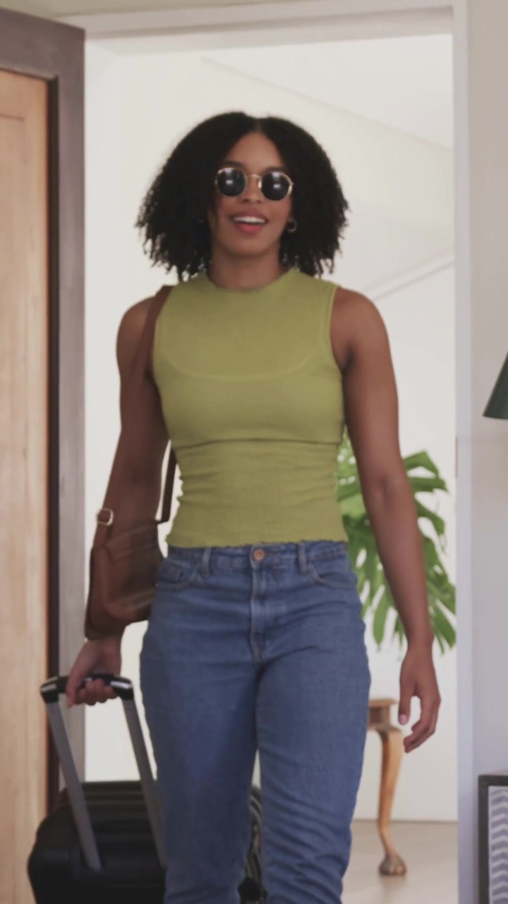 Donna che entra e sistema il manico telescopico di una valigia alla porta, con un top verde e jeans, mentre tiene una borsa.
