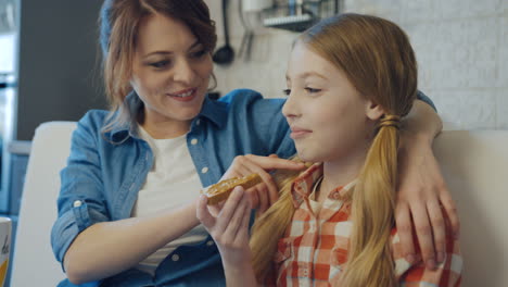 Close-up-of-the-pretty-teen-girl-biting-and-chewing-a-bread-with-peanut-butter-close-to-her-smiling-mother.-Portrait-shot.-Inside