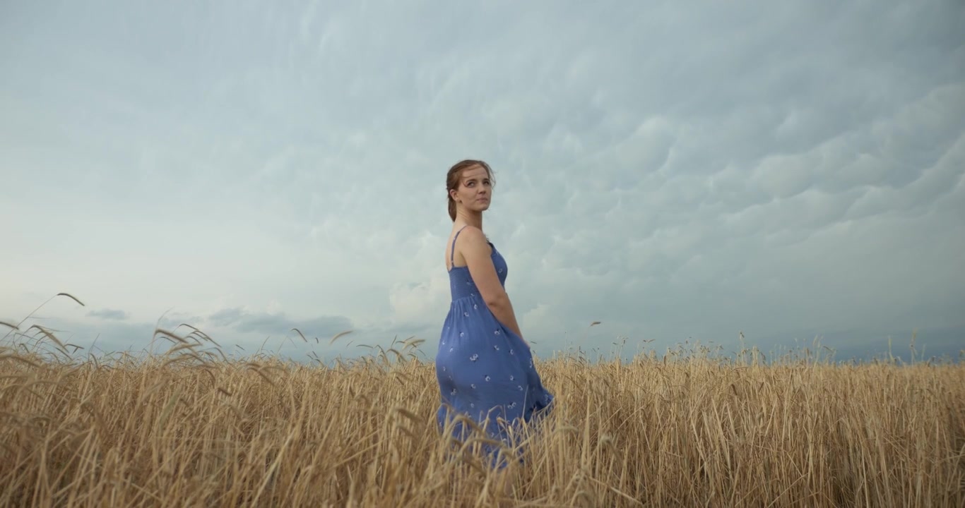 A young meditating, worshipping woman stands in the middle of a wheat field wearing a light blue dress with flowers, swaying gently in the breeze under a dramatic, stormy sky.