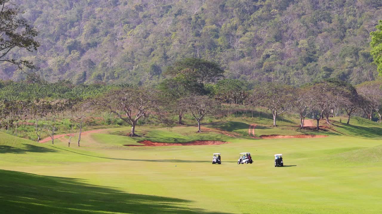 Golf carts move along green fairway with mountain backdrop, bright daylight, wide static shot