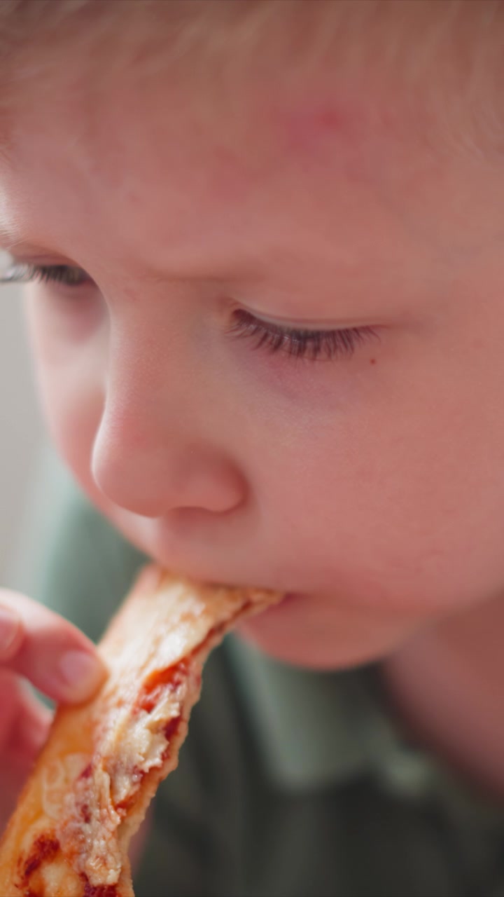 Close up of young boy eating pizza. child holds and takes bites from slice with focus on his facial expressions during consumption. Soft lighting highlights details in close proximity