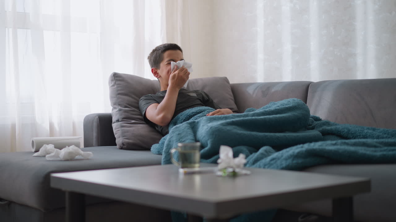 Sick little child sitting on couch sneezing into tissue, resting under blue blanket, with tissues on the table and a glass cup placed nearby, cozy home environment, sick child recovery