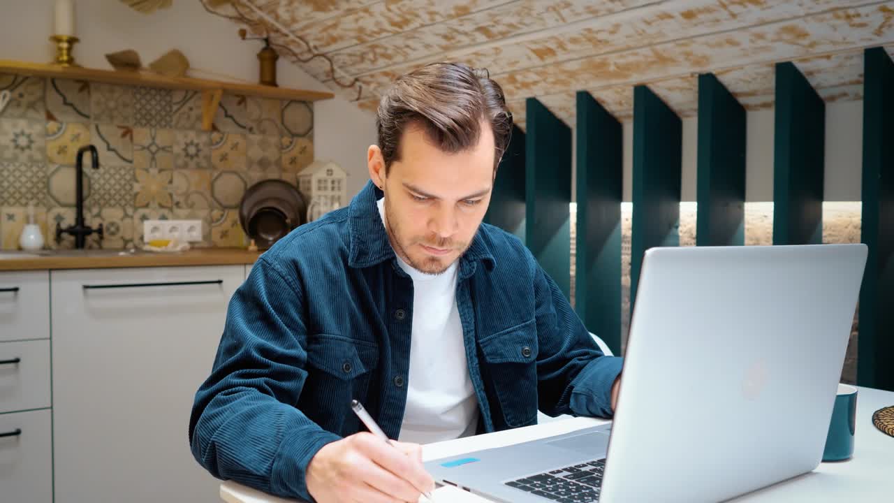 Frilanser jobber hjemmefra på kjøkkenet. Skriver et notat i en notatbok, ved siden av en laptop.