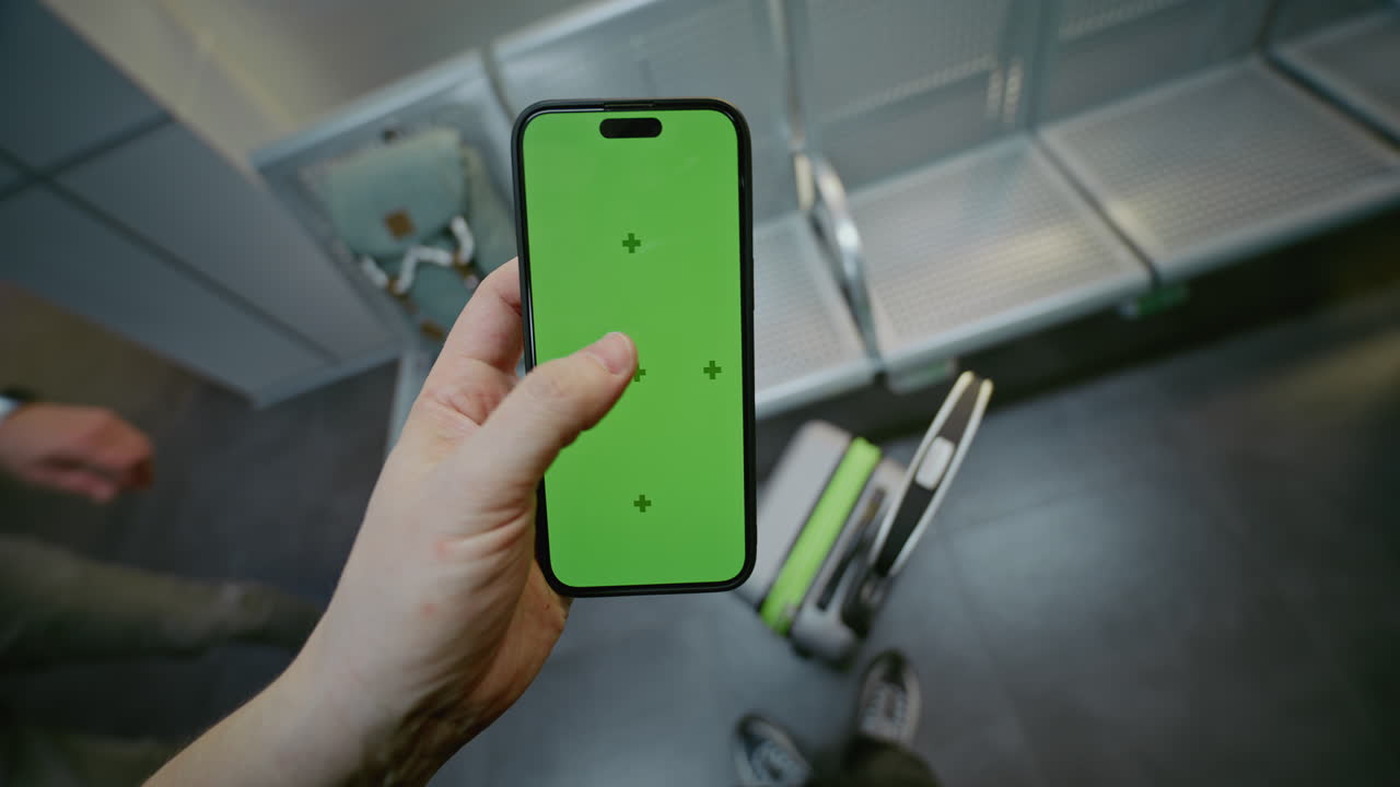 Person holding smartphone with green screen in an airport or train station waiting area.