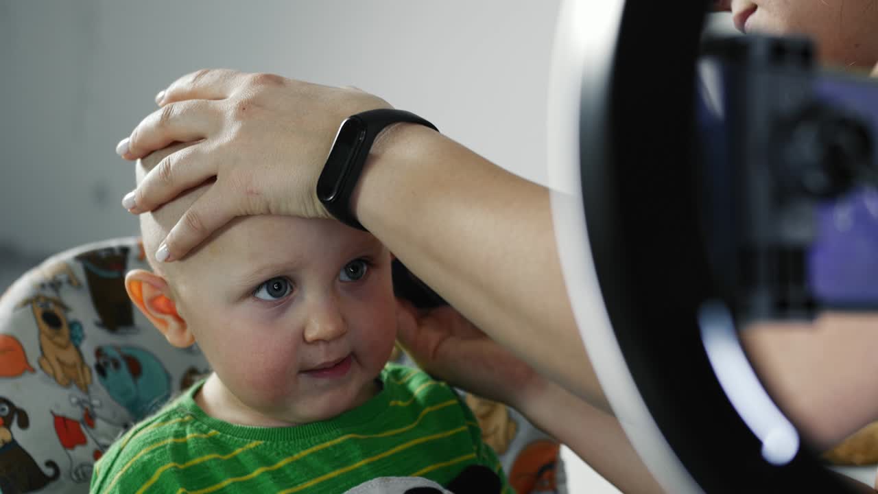 Mãe cortando o cabelo do filho pequeno com uma máquina de cortar cabelo na cozinha.